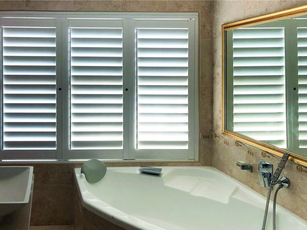 An image of a bathroom that showcases a corner bathtub, white aluminium shutters and a gold-framed mirror