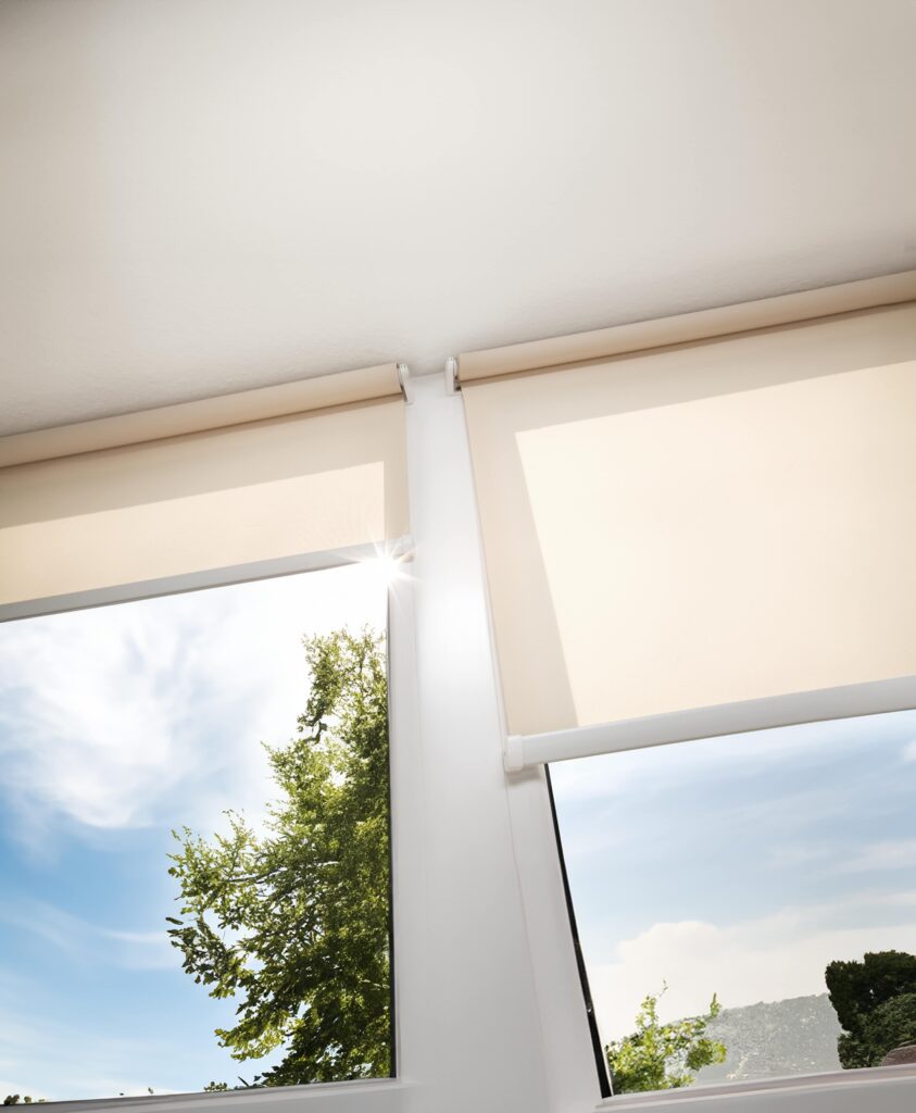 Upward facing view of windows with creme coloured roller blinds, open on both windows. A view of trees outside can be seen through the windows.