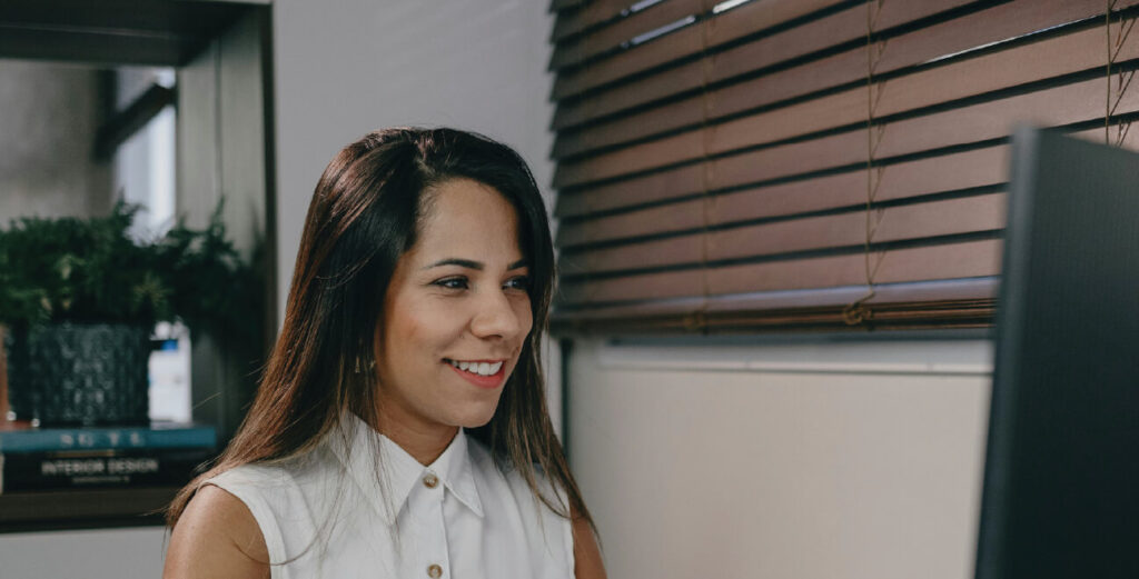 Lady smiling & staring at a computer screen, with dark brown wooden blinds to her side.