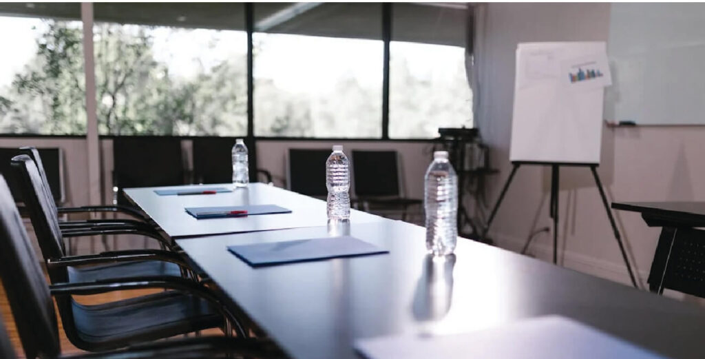 Long boardroom desk with windows at the back of the room with blinds.
